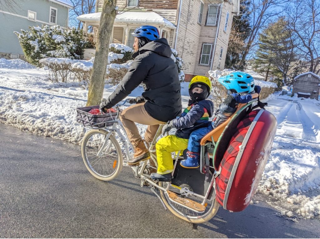 A father transports two children and a snow tube on a bicycle